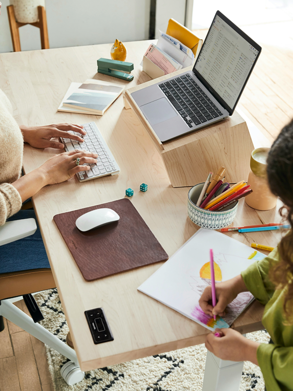 Eine Frau sitzt an einem Schreibtscih vor einem Laptop, daneben zeichnet ein Mädchen auf einem Malblock.