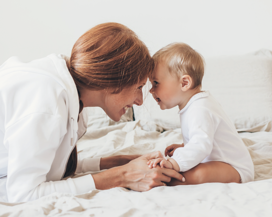 Eine Frau liegt lächelnd vor einem sitzenden Baby auf einem Bett.
