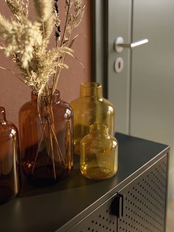 Nahaufnahme von gelben und braunen Glasvasen auf einer schwarzen Kommode vor einer terracottafarbenen Wand, im Hintergrund eine graue Tür