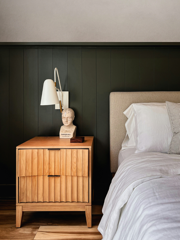 Graues Polsterbett und Beistelltisch aus hellem Holz vor einer dunkelgrünen Holzwand-Verkleidung