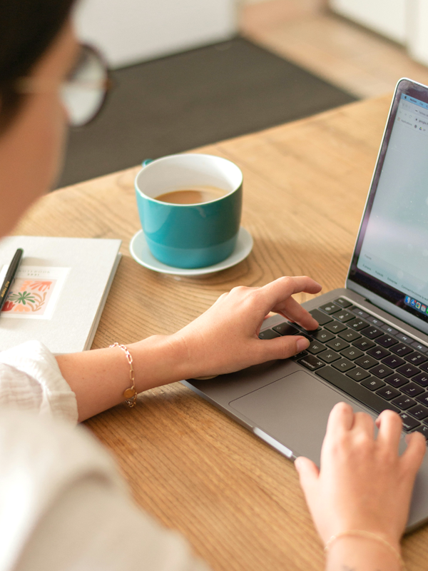 Eine Frau sitzt vor einem Laptop, neben einer Kaffeetasse und einem Notizbuch.