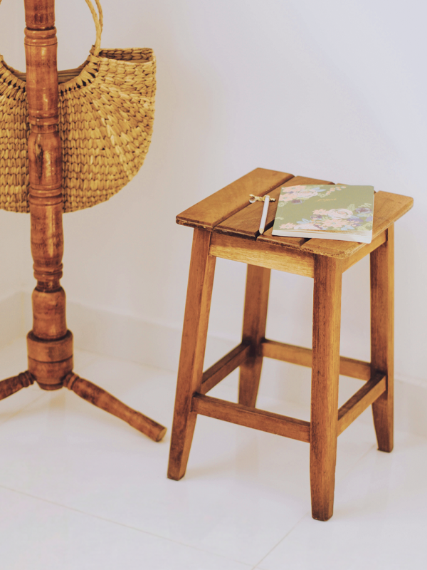 ein alter Holzhocker, auf dem ein Notizbuch liegt, daneben der Fuß eines alten Mantelständers aus Holz, an dem eine Korbtasche hängt