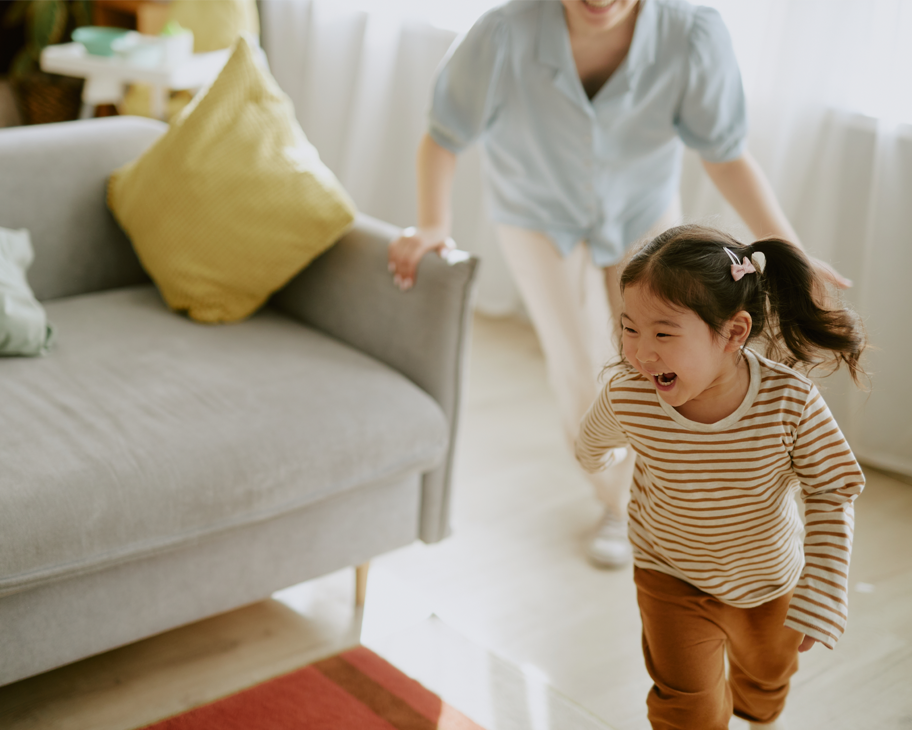 Ein Kleinkind läuft lachend mit einer Frau um ein graues Sofa herum.