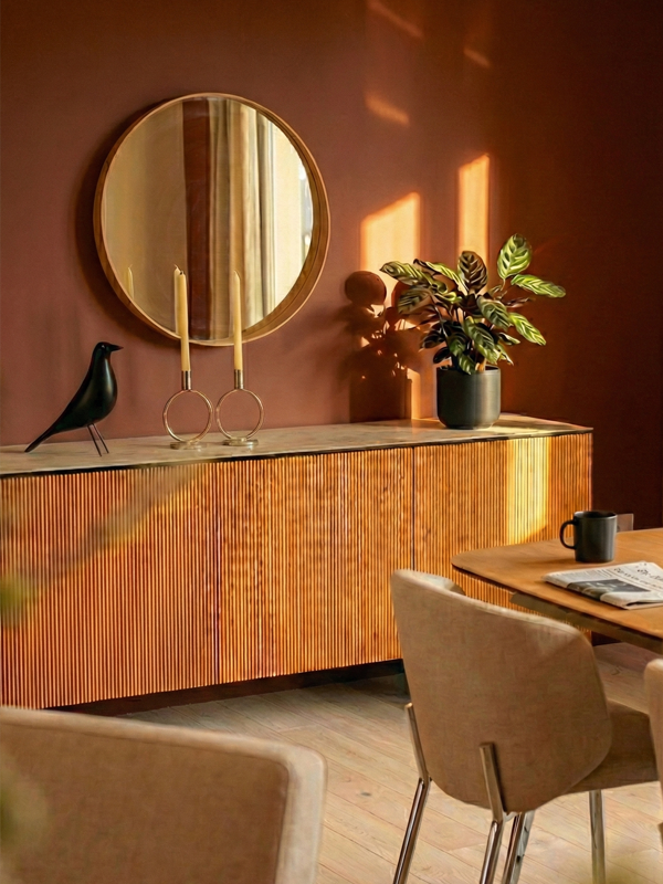 Ein Sideboard aus Holz mit Vogelfigur, Kerzenständern und Grünpflanze vor einer terracottafarbenen Wand mit rundem Spiegel.