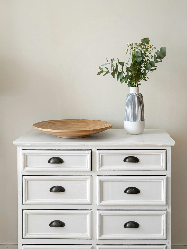 Eine weiße Landhauskommode mit einem Holzteller und einer Vase vor einer grauen Wand.