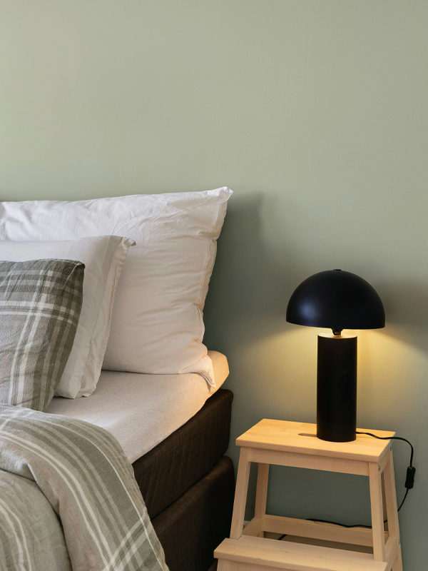 Ein Boxspringbett vor einer salbeigrünen Wand, daneben ein Holzhocker mit einer schwarzen Nachttischlampe.