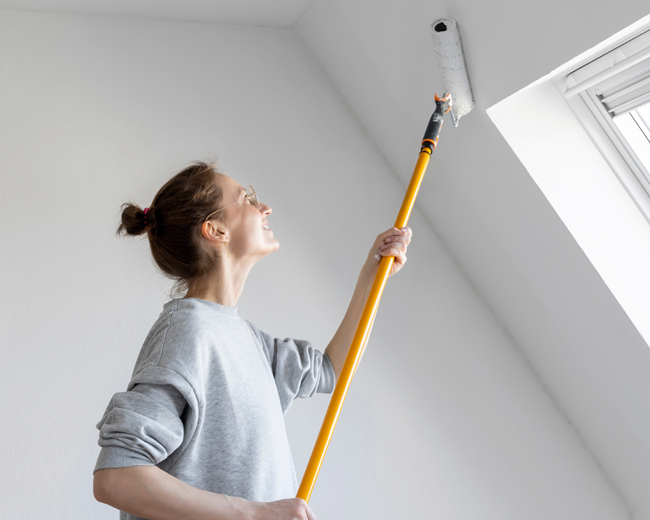 Eine junge Frau streicht mit einer Teleskop-Farbrolle eine Dachschräge weiß.
